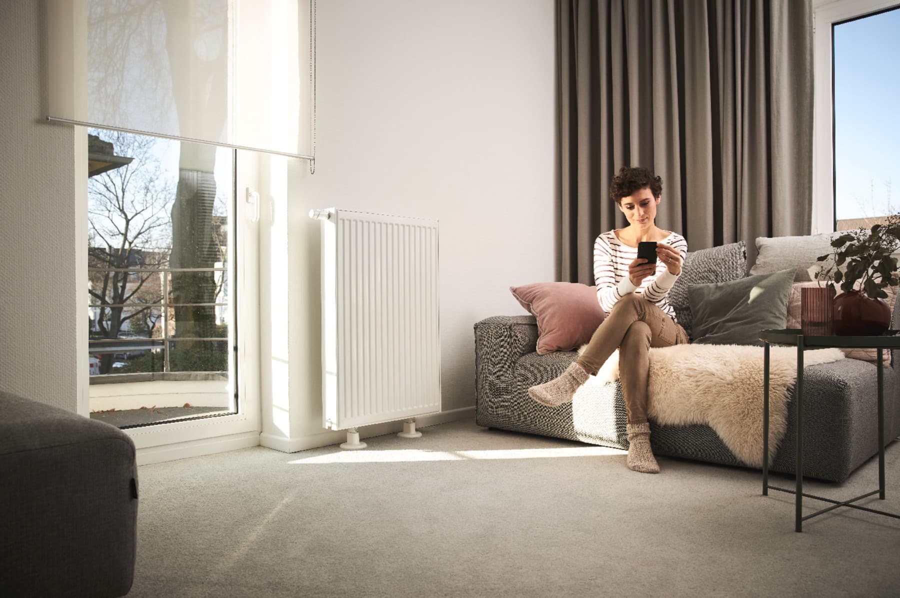 Kvinde sidder på sofaen ved siden af radiatoren og kigger på sin mobiltelefon