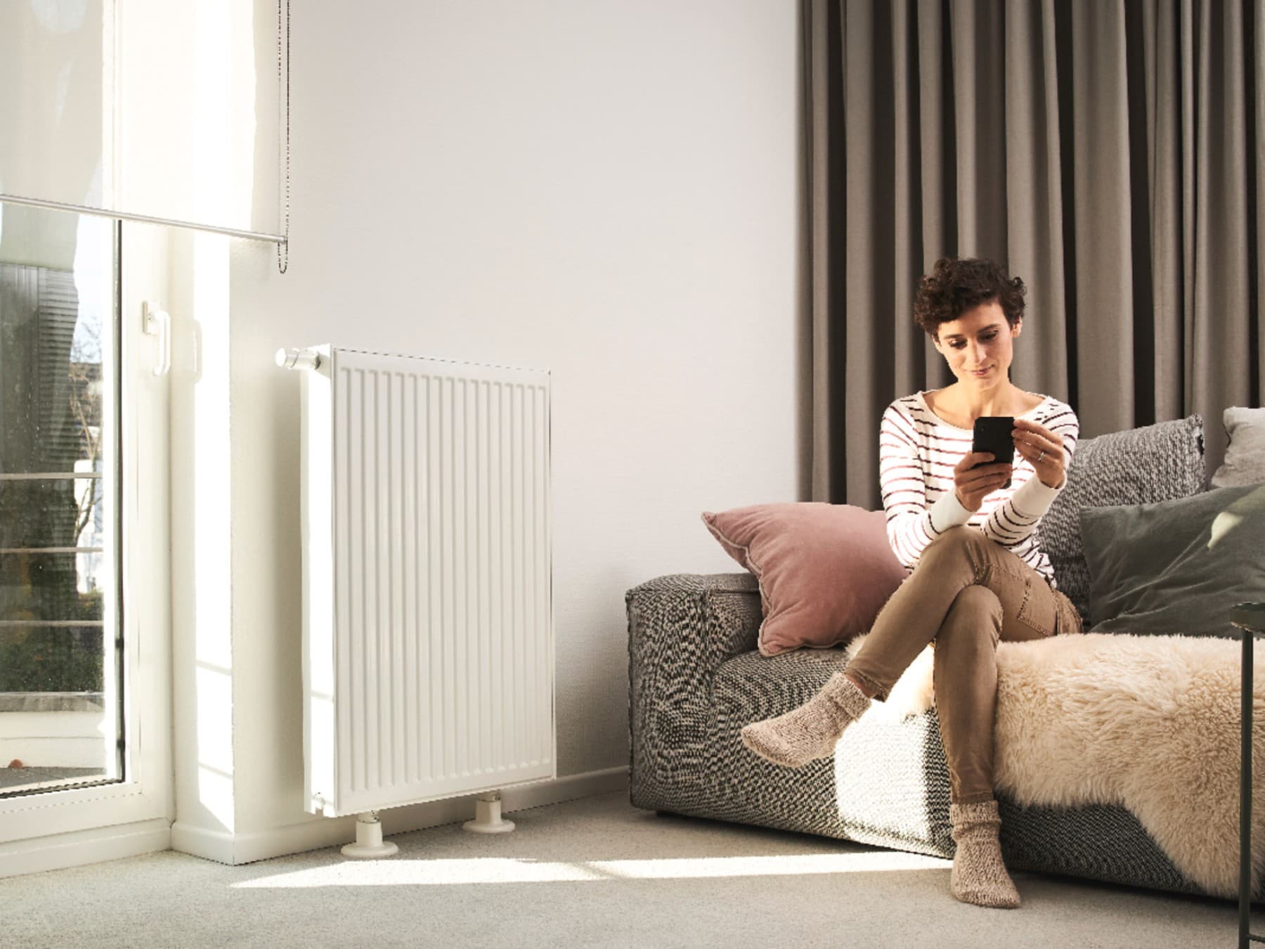 Kvinde sidder på sofaen og bruger en smartphone. Sofaen står foran et vindue og ved siden af en radiator.