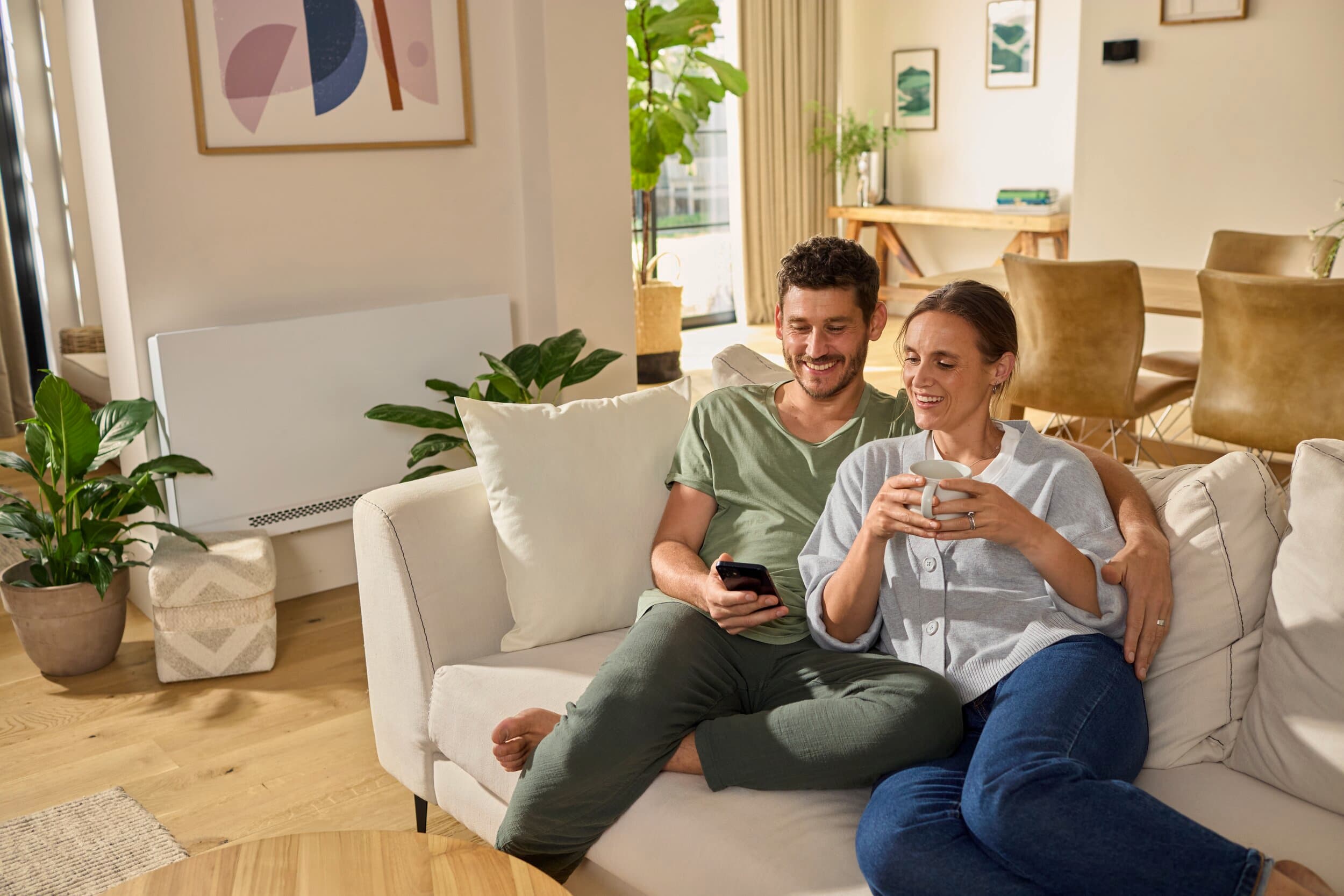 Et par, der slapper af på sofaen med en aroVAIR plus-ventilator-konvektor i baggrunden